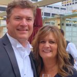 A candid outdoor photo of Pat Finn and a woman standing close together and smiling at the camera. Pat is dressed in a formal black suit with a white tuxedo shirt. They appear to be at an upscale rooftop venue with city buildings visible in the background.