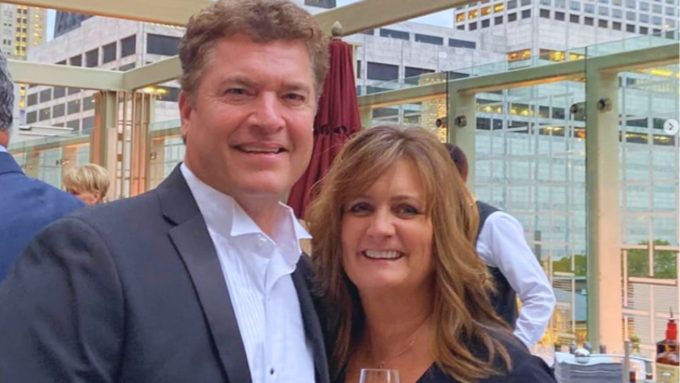 A candid outdoor photo of Pat Finn and a woman standing close together and smiling at the camera. Pat is dressed in a formal black suit with a white tuxedo shirt. They appear to be at an upscale rooftop venue with city buildings visible in the background.