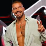 A smiling Ricky Martin posing on a red carpet, wearing an unbuttoned light green shirt that reveals his tattooed chest. He is wearing multiple gold necklaces with green pendants and holding up a peace sign while standing in front of bright stage lights.