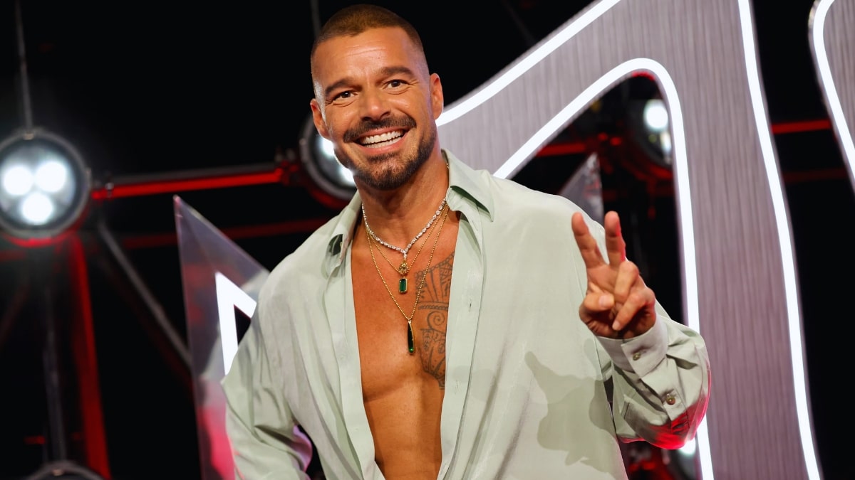 A smiling Ricky Martin posing on a red carpet, wearing an unbuttoned light green shirt that reveals his tattooed chest. He is wearing multiple gold necklaces with green pendants and holding up a peace sign while standing in front of bright stage lights.
