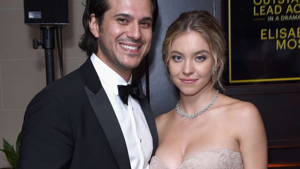 A red carpet photo of Sydney Sweeney and her partner, Jonathan Davino. Davino, on the left, is smiling and wearing a classic black tuxedo with a bow tie. Sweeney, on the right, wears a lace, champagne-colored strapless dress and a sparkling diamond necklace.