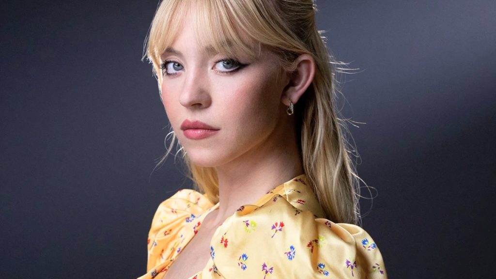 A close-up portrait of Sydney Sweeney looking directly at the camera. She has blonde hair styled with soft curtain bangs and a slight winged eyeliner look. She is wearing a yellow silk blouse featuring a delicate floral pattern of red, blue, and yellow flowers.