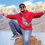 A full-body portrait of travel influencer Anunay Sood sitting on a rock in a vast mountain landscape. He is smiling with his arms outstretched, wearing a red hoodie, grey sweatpants, sunglasses, and a yellow cap. In the background, sun-drenched, rugged mountain peaks rise behind a calm, light blue high-altitude lake.