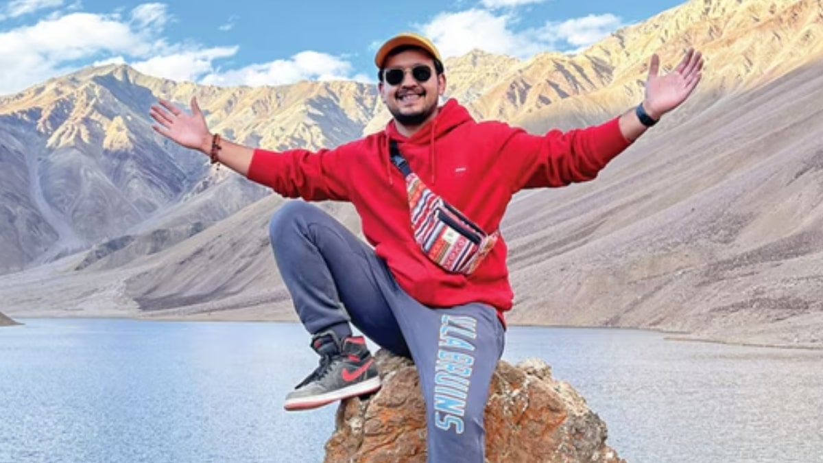 A full-body portrait of travel influencer Anunay Sood sitting on a rock in a vast mountain landscape. He is smiling with his arms outstretched, wearing a red hoodie, grey sweatpants, sunglasses, and a yellow cap. In the background, sun-drenched, rugged mountain peaks rise behind a calm, light blue high-altitude lake.