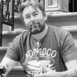 A black and white portrait of travel YouTuber Adam the Woo sitting on steps holding a camera, following news of his passing at age 51.