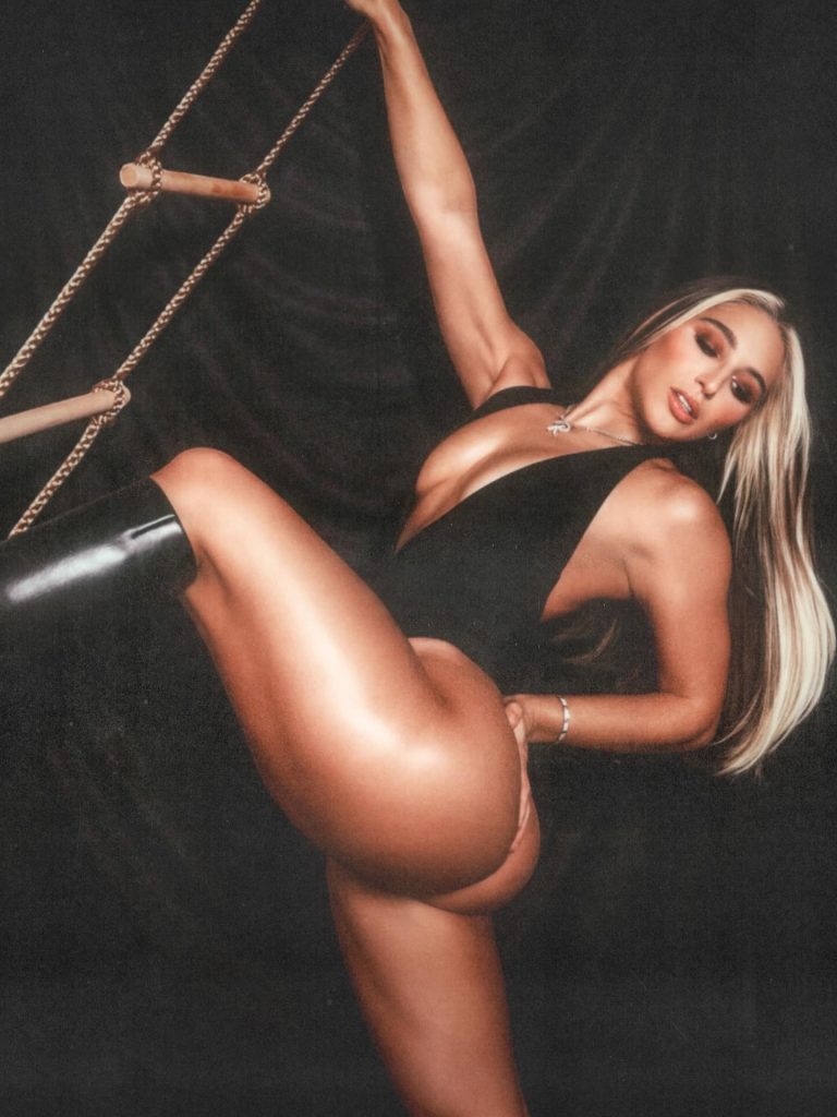A high-contrast studio photograph of Abella Danger posing dynamically on a rope ladder. She is wearing a plunging black bodysuit and black boots, set against a dark, textured background.