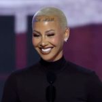Model and activist Amber Rose smiling on stage with her signature buzz cut and forehead tattoo during a public event.