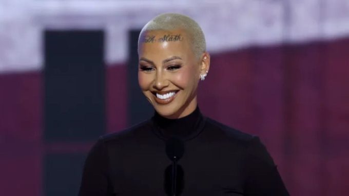 Model and activist Amber Rose smiling on stage with her signature buzz cut and forehead tattoo during a public event.
