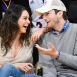 Ashton Kutcher and Mila Kunis (Talking) Ashton Kutcher and Mila Kunis are seen sitting courtside at a basketball game. Mila is laughing heartily while Ashton, wearing a white baseball cap and a grey sweater over a plaid shirt, gestures with his hand as they talk.
