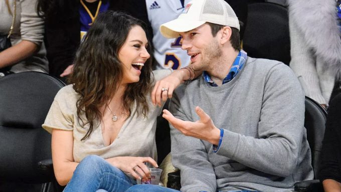 Ashton Kutcher and Mila Kunis (Talking) Ashton Kutcher and Mila Kunis are seen sitting courtside at a basketball game. Mila is laughing heartily while Ashton, wearing a white baseball cap and a grey sweater over a plaid shirt, gestures with his hand as they talk.
