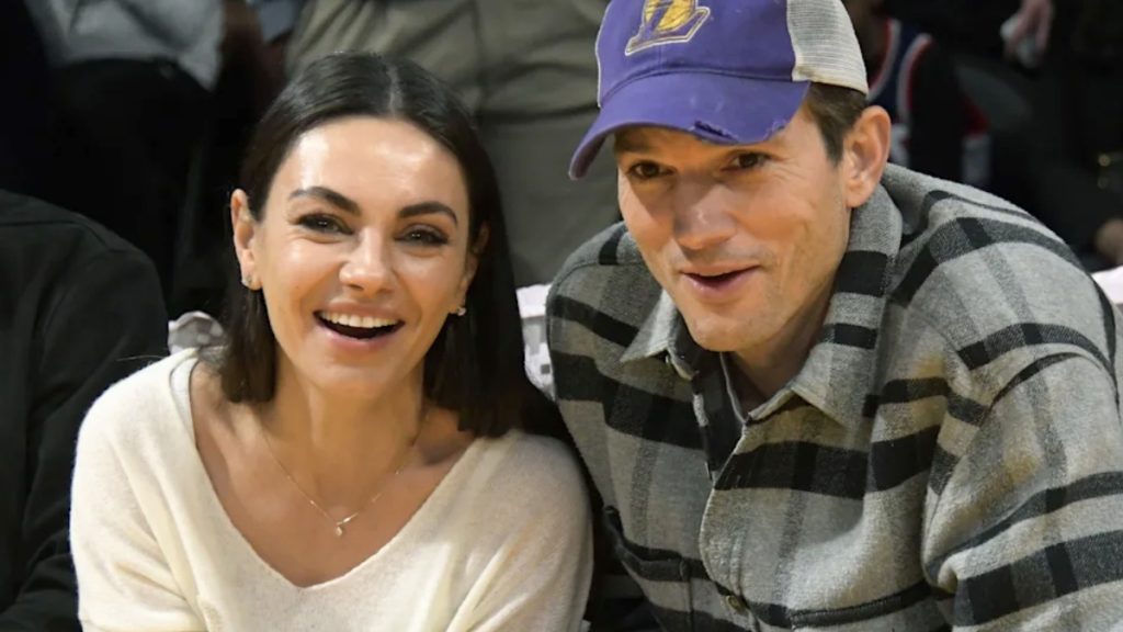 Ashton Kutcher and Mila Kunis (Smiling) Ashton Kutcher and Mila Kunis smile directly at the camera while attending an event. Mila is wearing a simple white V-neck sweater and a thin gold necklace, while Ashton wears a purple Los Angeles Lakers baseball cap and a grey and black flannel shirt.