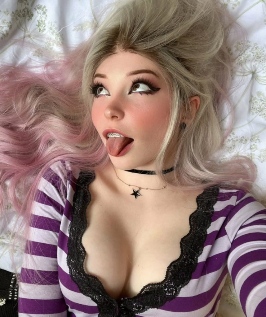 A close-up, high-angle photo of Belle Delphine lying on floral bedding. She is wearing a purple and white striped top with black lace trim and a black choker, featuring her signature playful expression and pink-tinted hair.