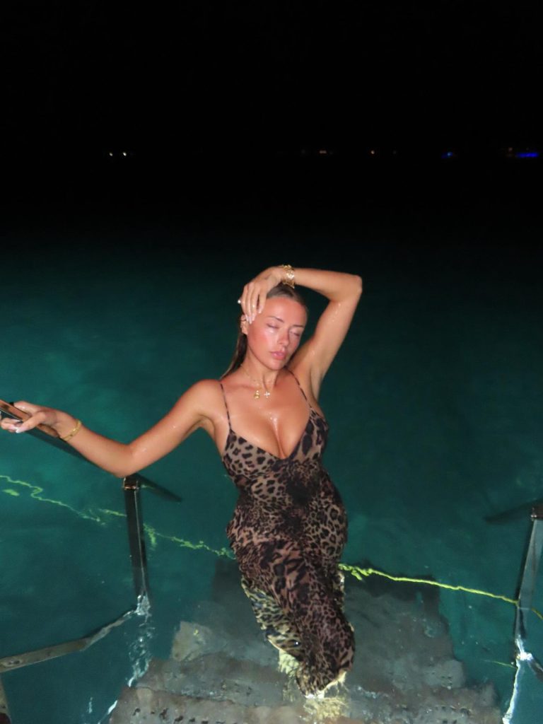 Corinna Kopf posing at night on the stairs of a boat or pier leading into glowing turquoise water. She is wearing a leopard-print maxi dress and has one arm raised to her head.