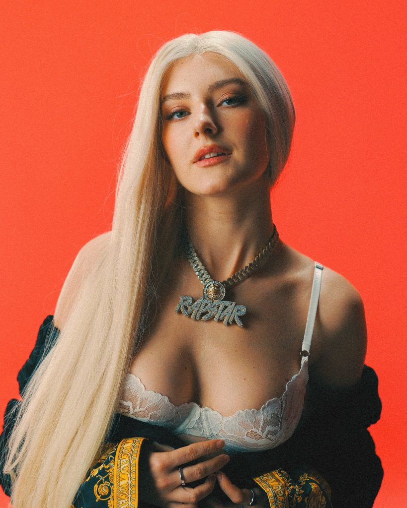 Eva Elfie with long blonde hair, wearing a white lace bra and a "RAPSTAR" diamond necklace against a solid red background.