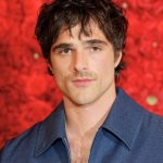 A red carpet style headshot of Jacob Elordi posing against a dense wall of red roses. He has dark, curly hair and a slight mustache, wearing a textured navy blue collared shirt that is partially unzipped. He is looking directly at the camera with a neutral expression.