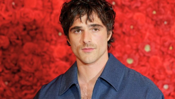 A red carpet style headshot of Jacob Elordi posing against a dense wall of red roses. He has dark, curly hair and a slight mustache, wearing a textured navy blue collared shirt that is partially unzipped. He is looking directly at the camera with a neutral expression.