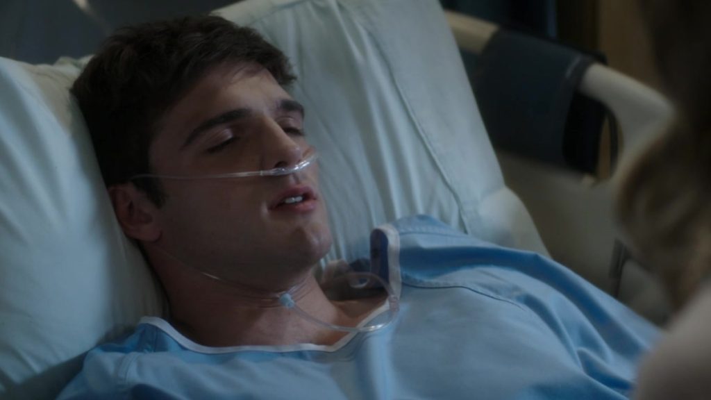 A close-up shot of actor Jacob Elordi lying in a hospital bed with his eyes closed. He is wearing a blue medical gown and has a clear nasal cannula providing oxygen. The lighting is dim, emphasizing a somber, clinical atmosphere.