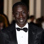A medium close-up of Khaby Lame, the famous TikTok creator, smiling while wearing a formal black tuxedo and bow tie. The image serves as a header for a news story regarding a significant financial deal for the creator.