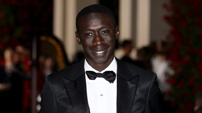 A medium close-up of Khaby Lame, the famous TikTok creator, smiling while wearing a formal black tuxedo and bow tie. The image serves as a header for a news story regarding a significant financial deal for the creator.