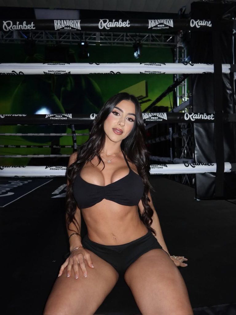 Lena The Plug sitting on the canvas of a boxing ring. She is wearing a black bikini-style top and black shorts, with "Rainbet" and "Brandrisk" branding visible on the ring ropes behind her.