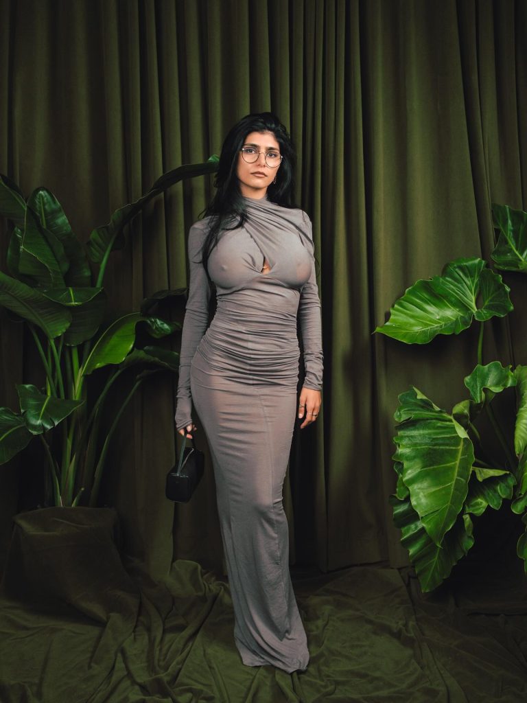 A full-length portrait of Mia Khalifa wearing a long-sleeved, floor-length grey dress and glasses. She is standing in front of dark green curtains flanked by large tropical potted plants.
