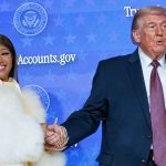 A medium-wide shot of Nicki Minaj and Donald Trump holding hands on a stage. Nicki Minaj is smiling brightly at the camera, wearing her signature white fur coat and long, neutral-toned manicured nails. Donald Trump is looking slightly to the side with an open-mouthed expression. The background is a blue digital screen with repeating Presidential Seals and text.