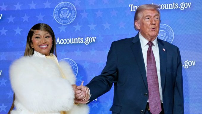 A medium-wide shot of Nicki Minaj and Donald Trump holding hands on a stage. Nicki Minaj is smiling brightly at the camera, wearing her signature white fur coat and long, neutral-toned manicured nails. Donald Trump is looking slightly to the side with an open-mouthed expression. The background is a blue digital screen with repeating Presidential Seals and text.