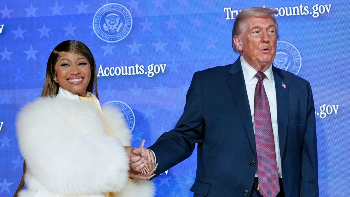A medium-wide shot of Nicki Minaj and Donald Trump holding hands on a stage. Nicki Minaj is smiling brightly at the camera, wearing her signature white fur coat and long, neutral-toned manicured nails. Donald Trump is looking slightly to the side with an open-mouthed expression. The background is a blue digital screen with repeating Presidential Seals and text.