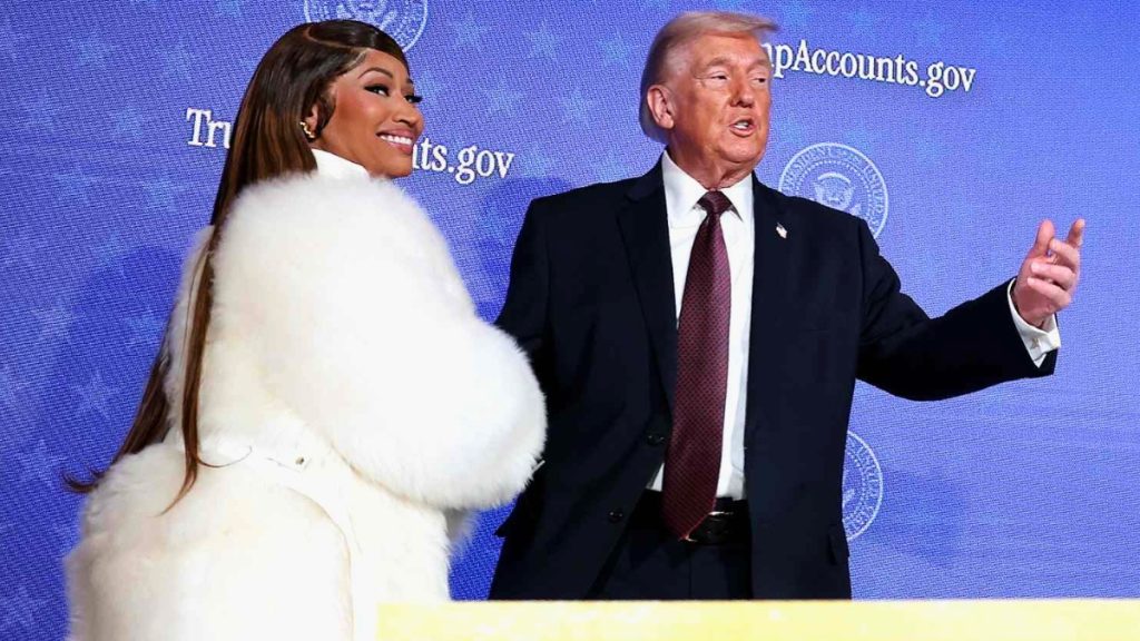 A side-angle shot of Nicki Minaj and Donald Trump on a stage in front of a blue backdrop featuring the Presidential Seal and a website URL. Nicki Minaj, seen mostly from the back showing her long brown hair and white fur coat, smiles over her shoulder toward Trump. Donald Trump is gesturing with his left hand while speaking into the distance.