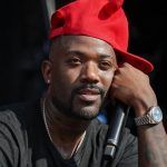 A close-up shot of Ray J outdoors, looking upward with a thoughtful expression while resting his chin on his hand. He is wearing a bright red baseball cap, a black t-shirt, and a silver wristwatch, while holding a black microphone.