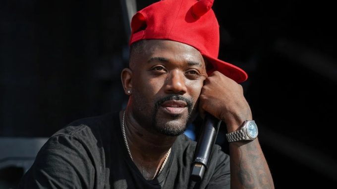 A close-up shot of Ray J outdoors, looking upward with a thoughtful expression while resting his chin on his hand. He is wearing a bright red baseball cap, a black t-shirt, and a silver wristwatch, while holding a black microphone.