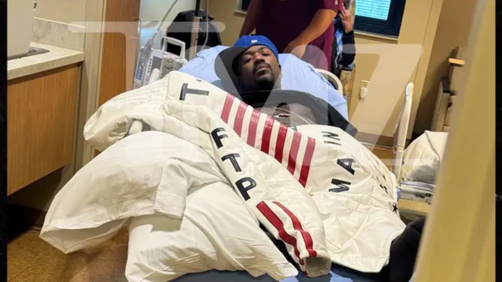 Ray J lies in a hospital bed wearing a blue baseball cap, partially covered by a white jacket with red stripes and the letters "FTP". A large, translucent "TMZ" watermark is layered over the center of the image, which shows medical equipment in the background and a person in purple scrubs standing nearby.