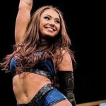 Wrestler Skye Blue smiling with one arm raised, wearing a blue and black glittery athletic top and arm guard against a dark background.