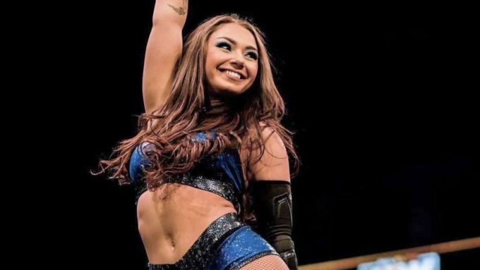 Wrestler Skye Blue smiling with one arm raised, wearing a blue and black glittery athletic top and arm guard against a dark background.