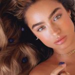 A high-angle, close-up portrait of Sommer Ray lying on a white, sheer fabric. She has long, wavy light brown hair spread out around her, accented with small purple berries. She is looking directly at the camera with a neutral expression, wearing a gold nameplate necklace that reads "IMARAÏS" and holding a single purple berry near her lips.