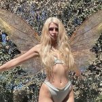 Sunny Rayyxo posing outdoors wearing large iridescent fairy wings and a white bikini against a lush green forest background.