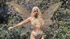 Sunny Rayyxo posing outdoors wearing large iridescent fairy wings and a white bikini against a lush green forest background.