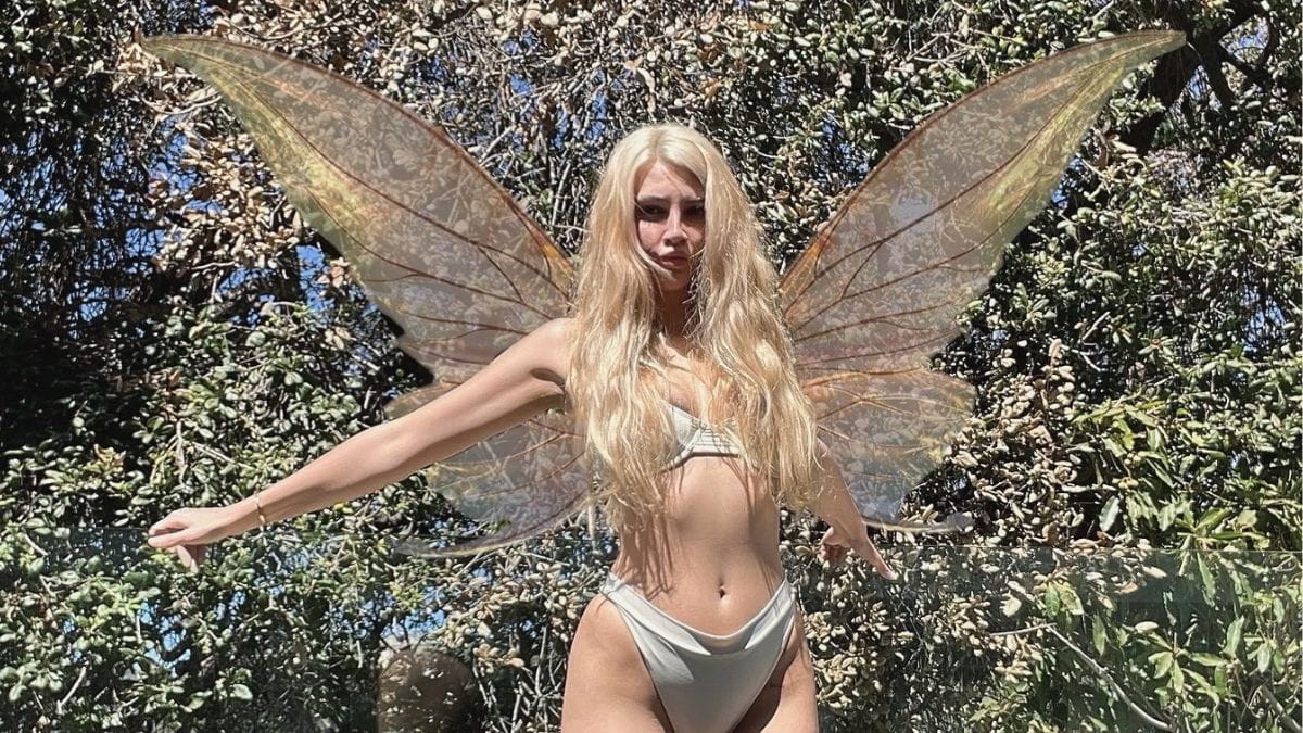 Sunny Rayyxo posing outdoors wearing large iridescent fairy wings and a white bikini against a lush green forest background.