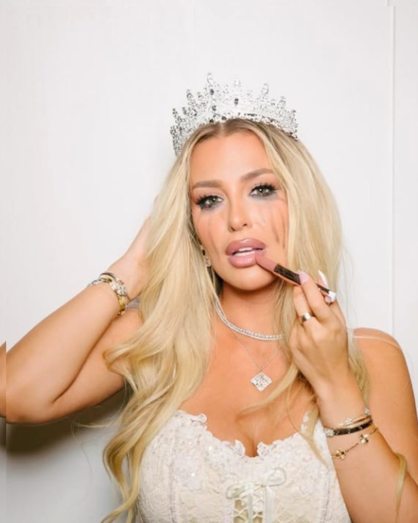 YouTuber Tana Mongeau wearing a crystal tiara and white lace corset while applying lipstick with smudged mascara makeup.