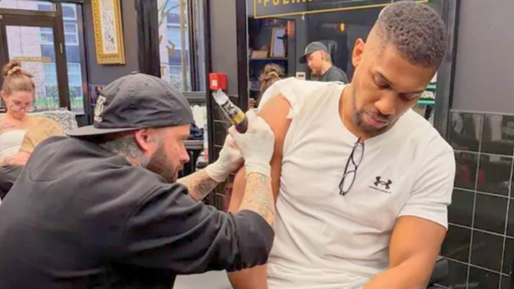 A medium shot inside a tattoo parlor showing Anthony Joshua sitting in a chair, wearing a white Under Armour t-shirt. He is looking down as a male tattoo artist with a black cap and tattooed arms works on Joshua’s right shoulder using a tattoo machine. The atmosphere appears somber and focused.
