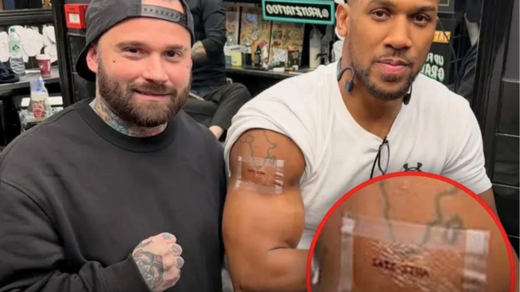 A medium shot of Anthony Joshua posing with his tattoo artist. Joshua is wearing a white t-shirt with the right sleeve pushed up to reveal a fresh tattoo on his bicep. The tattoo features an outline of the continent of Africa with text underneath. A red circular inset provides a zoomed-in view of the tattoo, which appears to read "LATE SIRA."