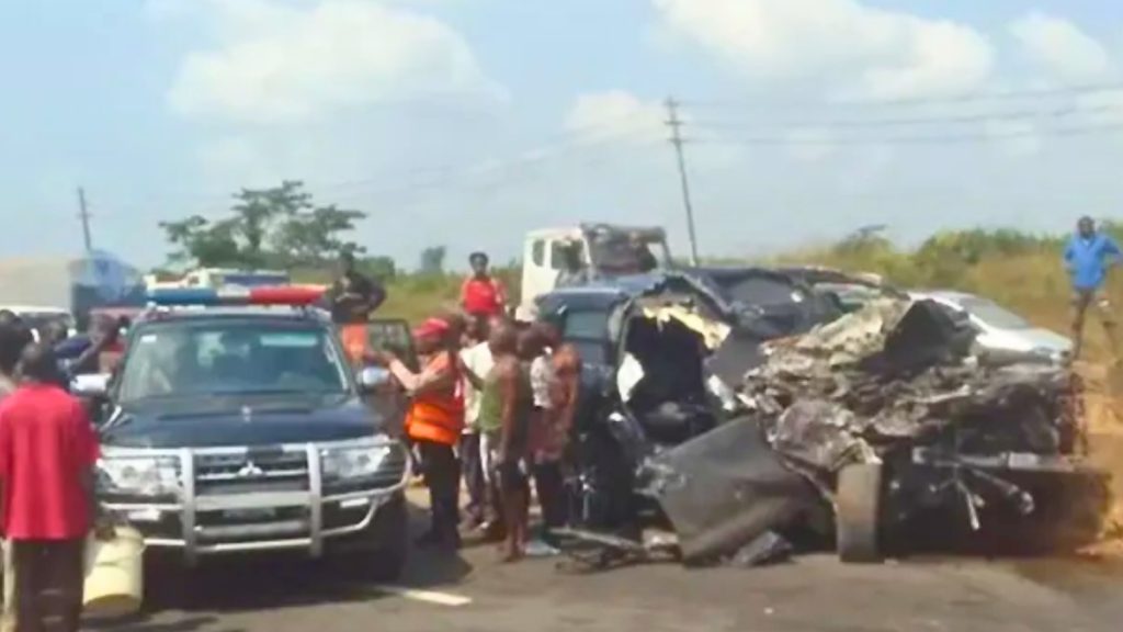 A wide shot of a tragic car accident on a sunlit road. A black SUV is severely crushed and mangled beyond recognition on the right side of the frame. To the left, a dark Mitsubishi police vehicle with emergency lights is parked. Several onlookers and emergency responders in orange vests are gathered around the wreckage.