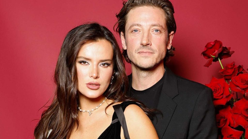 A romantic portrait of Bella Thorne with fiancé Mark Emms, posing against a deep red background with a bouquet of red roses.