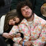 A photograph of Selena Gomez and Benny Blanco sitting together at a public event. Gomez is leaning her head on Blanco’s shoulder while looking down at her phone. Blanco, wearing a white puffer jacket with a pink and orange floral pattern, is looking upward. A small circular inset in the top right corner shows a close-up of a person's bare, dirty feet resting on a grey sofa.