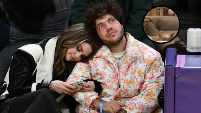 A photograph of Selena Gomez and Benny Blanco sitting together at a public event. Gomez is leaning her head on Blanco’s shoulder while looking down at her phone. Blanco, wearing a white puffer jacket with a pink and orange floral pattern, is looking upward. A small circular inset in the top right corner shows a close-up of a person's bare, dirty feet resting on a grey sofa.