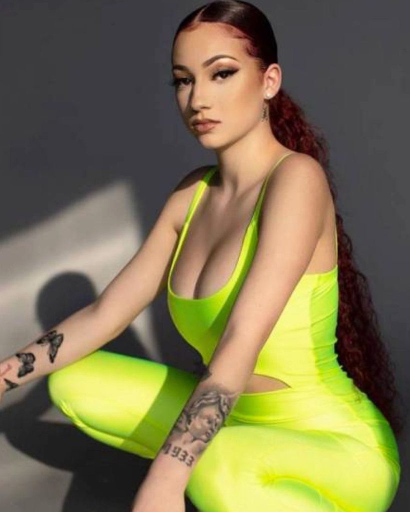 Bhad Bhabie wearing a neon yellow athletic bodysuit with visible arm tattoos and long wavy hair.