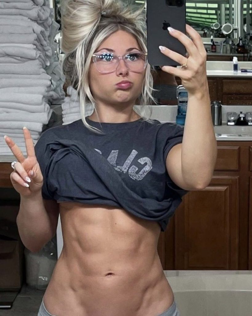 Carriejune Anne Bowlby, fitness influencer and Mini Beast founder, showing her toned abs in a bathroom mirror selfie. She is wearing a grey cropped t-shirt, glasses, and has her hair in a messy bun, posing with a peace sign.