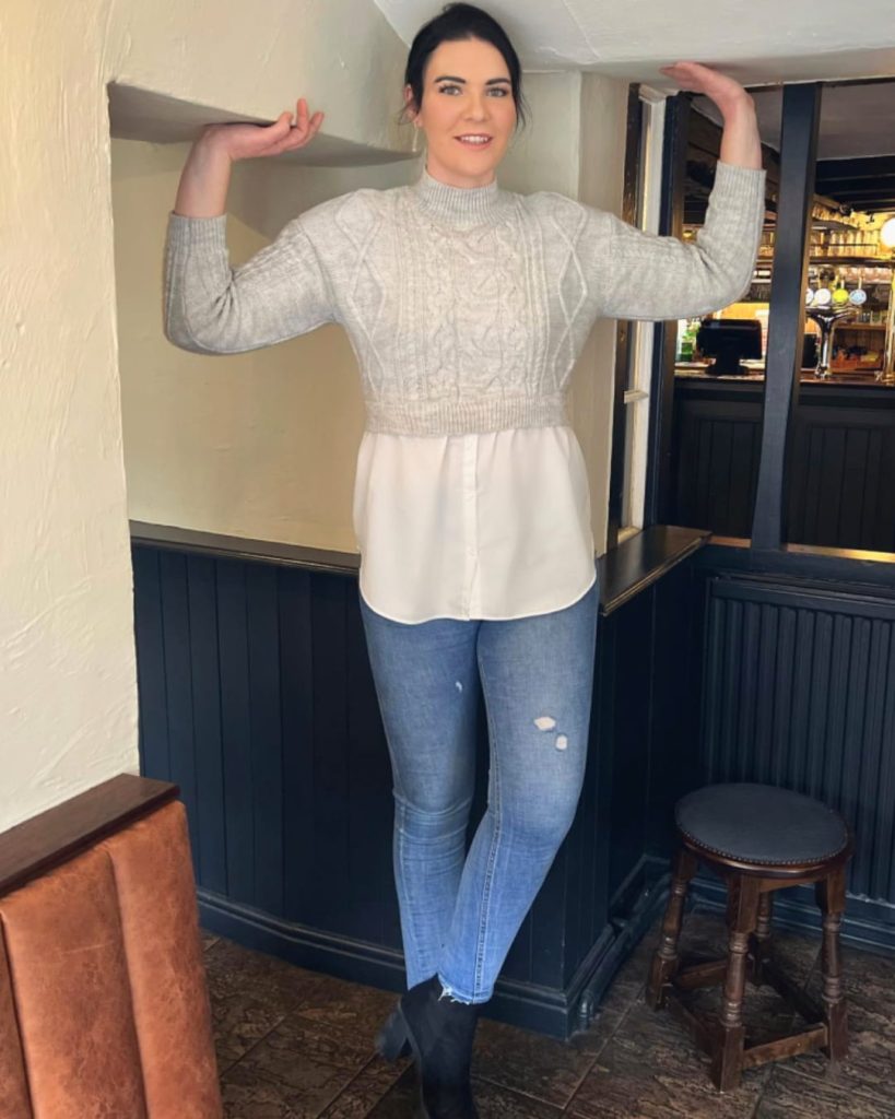 Tall influencer Charlie Mill posing in a pub, standing at full height and touching the ceiling to show stature, wearing a grey cable-knit sweater and blue jeans.