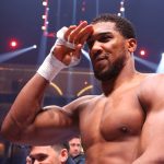 A high-resolution, medium shot of professional boxer Anthony Joshua in a boxing ring under bright arena lights. He is shirtless, showing his muscular physique, with his right hand wrapped in white boxing tape and raised to his forehead in a salute. The background is blurred, showing the tiered seating of a large arena.