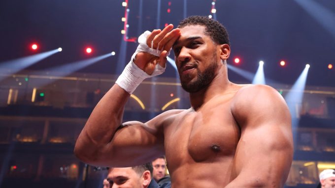 A high-resolution, medium shot of professional boxer Anthony Joshua in a boxing ring under bright arena lights. He is shirtless, showing his muscular physique, with his right hand wrapped in white boxing tape and raised to his forehead in a salute. The background is blurred, showing the tiered seating of a large arena.